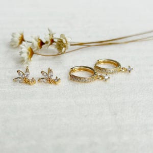 Puede incluir: Juego de joyas doradas sobre un fondo blanco. El conjunto incluye dos pares de pendientes: uno con diseño de hoja y otro con aro y un dije de estrella colgante. Los pendientes están adornados con pequeñas gemas transparentes.