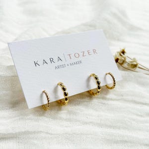 May include: A set of three gold-colored hoop earrings with different textures. One pair has a twisted rope design, another has a series of small beads, and the third has a textured surface. The earrings are displayed on a white card with the text "KARA TOZER ARTIST + MAKER".