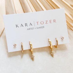 May include: A set of gold-toned earrings on a white display card labeled KARA TOZER ARTIST + MAKER. The set includes two small plain hoop earrings, two hoop earrings with dangling cross charms encrusted with clear stones, and two small stud earrings.