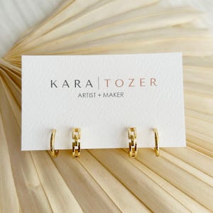 May include: A pair of gold hoop earrings with a textured chain-link design attached to a white display card. The card features the black and copper text KARA TOZER ARTIST + MAKER. The earrings are polished and have a reflective metallic finish.