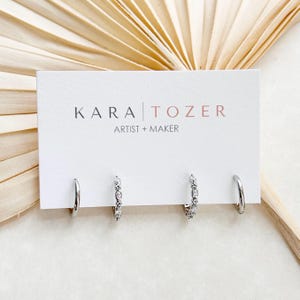 May include: Four silver-toned hoop earrings displayed on a white rectangular card with the text KARA TOZER ARTIST + MAKER. Two hoops are plain polished metal, while the other two feature a row of small, clear, sparkling stones along the front.