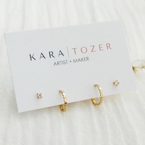 May include: A set of gold-colored earrings on a white card with the text "KARA TOZER ARTIST + MAKER". The set includes two pairs of stud earrings and two pairs of hoop earrings. One pair of studs has a square design, and the other has a flower design.