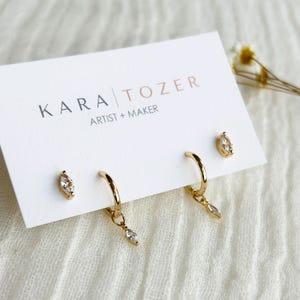 May include: Gold-toned jewelry set featuring two pairs of earrings. One pair is small studs with a marquise-cut clear stone. The other pair are small hoop earrings with a dangling marquise-cut clear stone. The earrings are displayed on a white card with the text "KARA TOZER ARTIST + MAKER".