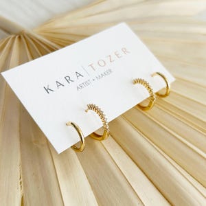 May include: Four gold hoop earrings attached to a white display card with the text KARA TOZER ARTIST + MAKER. Two hoops are plain gold, and two hoops feature a row of small clear stones along the outer edge. The earrings are arranged in pairs.