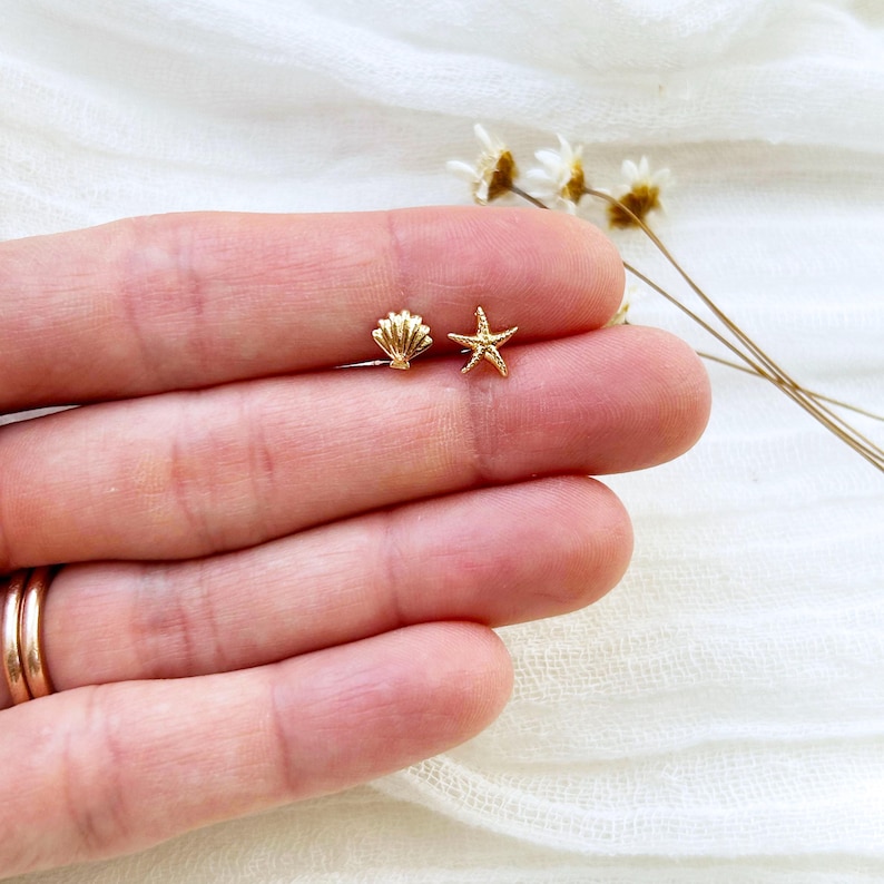 May include: A pair of gold-colored stud earrings. One earring is shaped like a seashell, and the other is a starfish. The earrings are small and delicate, perfect for everyday wear. The earrings are displayed on a person's hand.