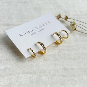 May include: A set of gold-colored hoop earrings, including two pairs of simple hoops and two pairs with a chain design. The earrings are displayed on a white card with the text "KARA TOZER ARTIST + MAKER".
