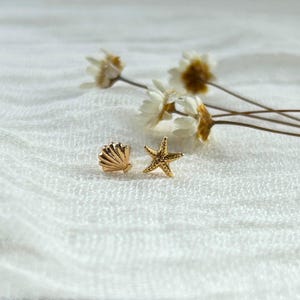 May include: A pair of gold-colored stud earrings. One earring is shaped like a seashell, and the other is a starfish. The earrings are on a white, textured surface with dried flowers in the background.
