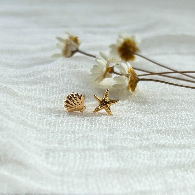May include: A pair of gold-colored stud earrings. One earring is shaped like a seashell, and the other is a starfish. The earrings are on a white, textured surface with dried flowers in the background.