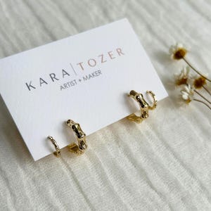 May include: Three pairs of gold-colored hoop earrings with a bamboo design are displayed on a white card. The card has the text "KARA TOZER ARTIST + MAKER". The earrings are on a white surface with a few small, dried flowers.
