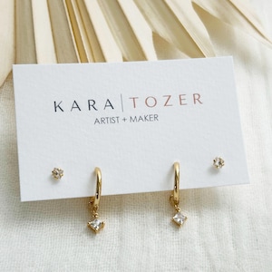 May include: A set of gold-toned jewelry on a white display card labeled KARA TOZER ARTIST + MAKER. The set includes two small stud earrings with clear stones and two gold hoop earrings, each featuring a dangling square-cut clear stone charm.