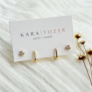 May include: A white display card with the black and copper text KARA TOZER ARTIST + MAKER features two gold hoop earrings and two gold stud earrings with three small clear stones arranged in a cluster on each stud earring.