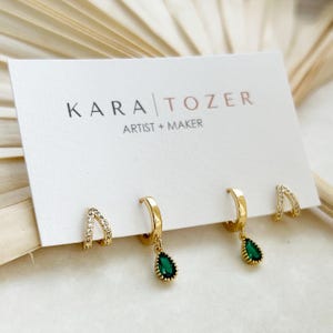 May include: Four gold-tone hoop earrings displayed on a white card labeled KARA TOZER ARTIST + MAKER. Two earrings are plain hoops with dangling green teardrop-shaped stones, and two are small hoops encrusted with clear stones in a V-shape design.