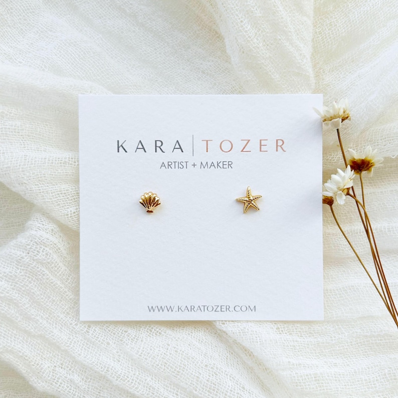 May include: A pair of gold-colored stud earrings, one shaped like a seashell and the other a starfish, displayed on a white card. The card has the text "KARA TOZER ARTIST + MAKER" and the website address. The background is a white, textured fabric.
