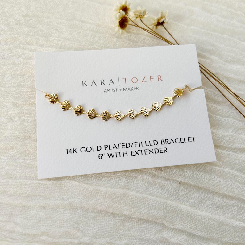 May include: A gold plated or filled bracelet with a delicate seashell design. The bracelet is 6 inches long with an extender. The bracelet is displayed on a white card with the text "KARA TOZER ARTIST + MAKER 14K GOLD PLATED/FILLED BRACELET 6" WITH EXTENDER"