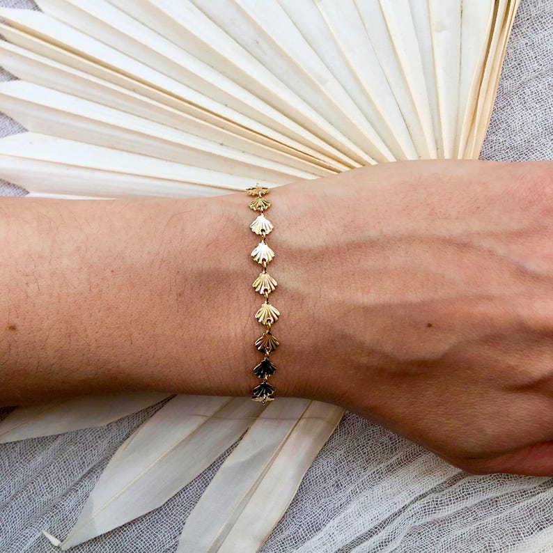 May include: A gold bracelet with a delicate chain and small, scallop-shaped charms. The bracelet is on a person's wrist.