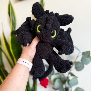 Puede incluir: Un peluche de dragón negro tejido a ganchillo, con grandes ojos verdes y una punta de cola roja. El dragón tiene alas y está hecho de un hilo suave y texturizado. Un peluche hecho a mano, perfecto para una habitación infantil o como regalo.