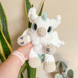 May include: A handmade white crochet griffin plush toy with light blue accents. The plush has large black eyes with blue rims, a tan beak, and tan feet with purple accents. The griffin is held in front of a plant.