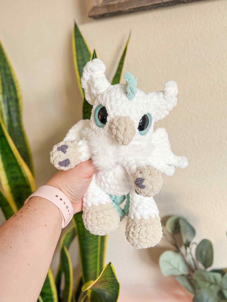 May include: A handmade white crochet griffin plush toy with light blue accents. The plush has large black eyes with blue rims, a tan beak, and tan feet with purple accents. The griffin is held in front of a plant.
