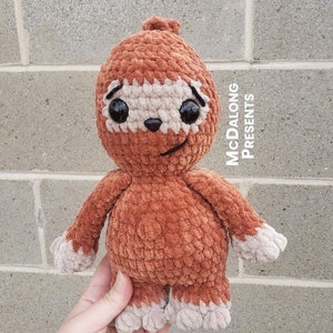 May include: A crocheted sloth toy with brown fur and a beige face. The sloth has large black eyes and a small, beige nose. The toy is held in a hand against a gray brick wall. The text "McDalong Presents" is visible in the upper right corner of the image.