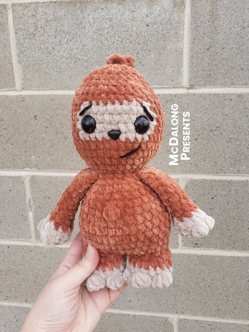 May include: A crocheted sloth toy with brown fur and a beige face. The sloth has large black eyes and a small, beige nose. The toy is held in a hand against a gray brick wall. The text "McDalong Presents" is visible in the upper right corner of the image.