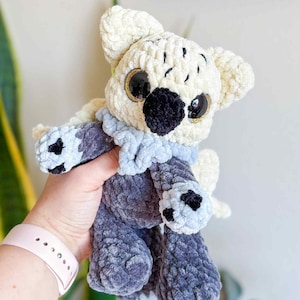 May include: A handmade plush toy in the shape of a koala bear. The koala is made of soft, cream-colored, and gray yarn. It has large, black eyes with gold accents, a black nose, and a light blue scarf.