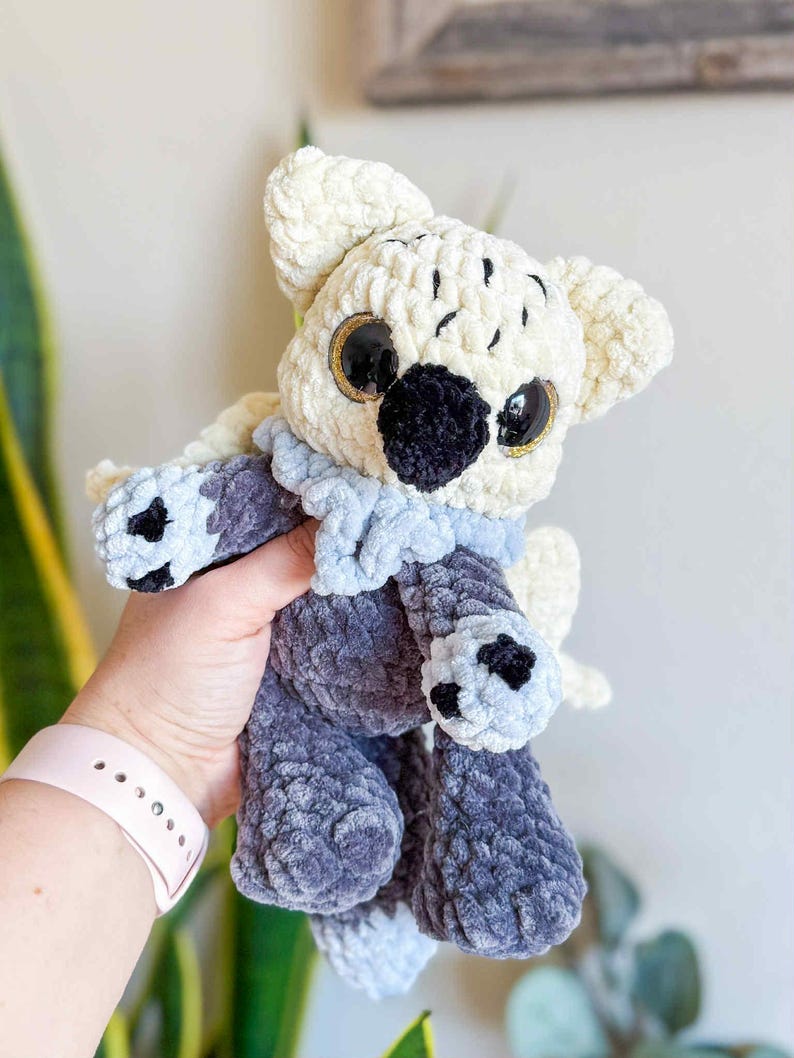 May include: A handmade plush toy in the shape of a koala bear. The koala is made of soft, cream-colored, and gray yarn. It has large, black eyes with gold accents, a black nose, and a light blue scarf.