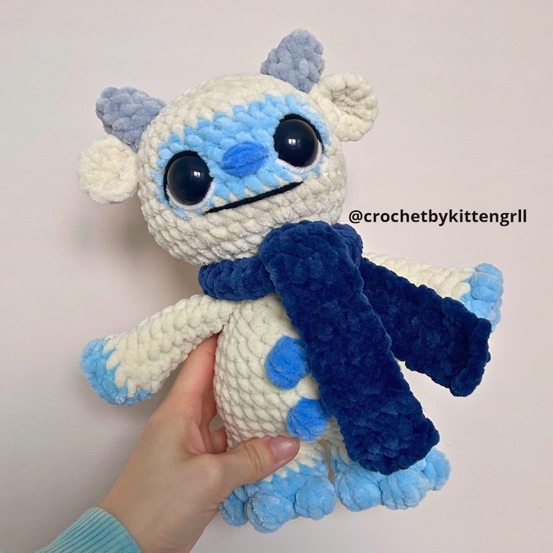 May include: A white and blue crocheted plush toy with large black eyes and a blue scarf. The toy has a textured, fuzzy appearance and is holding a blue scarf.