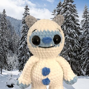 May include: A white crocheted plush toy with blue accents resembling a yeti or abominable snowman. The toy has large black eyes and a smiling mouth. The toy is standing in a snowy winter scene.