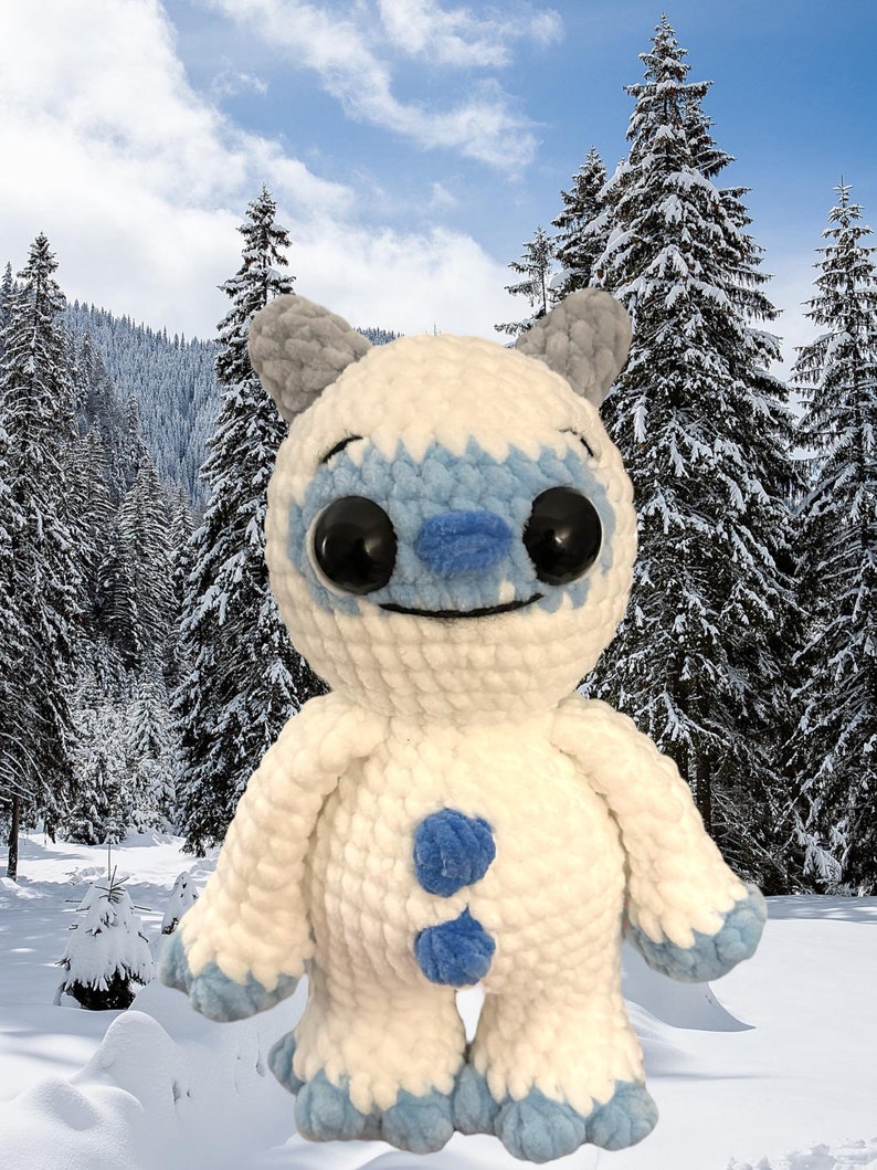 May include: A white crocheted plush toy with blue accents resembling a yeti or abominable snowman. The toy has large black eyes and a smiling mouth. The toy is standing in a snowy winter scene.