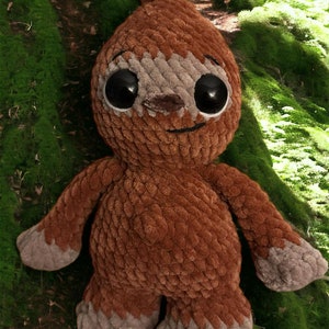 May include: A brown crocheted sloth plush toy with large black eyes and a smiling mouth. The toy is sitting on a bed of green moss.