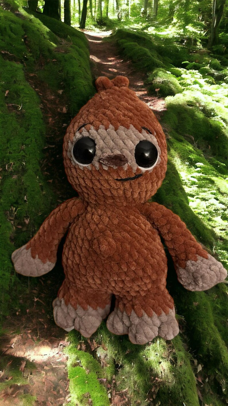 May include: A brown crocheted sloth plush toy with large black eyes and a smiling mouth. The toy is sitting on a bed of green moss.