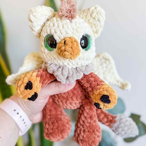 May include: A handmade crochet griffin plush toy. The plush has a cream-colored head with large green eyes, a tan beak, and a tuft of pink hair. The body is a mix of orange and brown, with small wings and black accents on the feet.