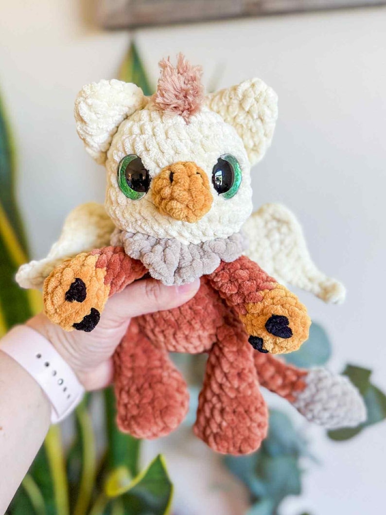 May include: A handmade crochet griffin plush toy. The plush has a cream-colored head with large green eyes, a tan beak, and a tuft of pink hair. The body is a mix of orange and brown, with small wings and black accents on the feet.