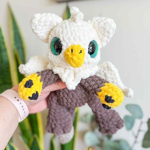 May include: A handmade crochet plush toy in the shape of a bird. The toy features a white head and wings, a yellow beak, and large green eyes. The body is brown with yellow and black accents on the feet. The plush is held by a person.