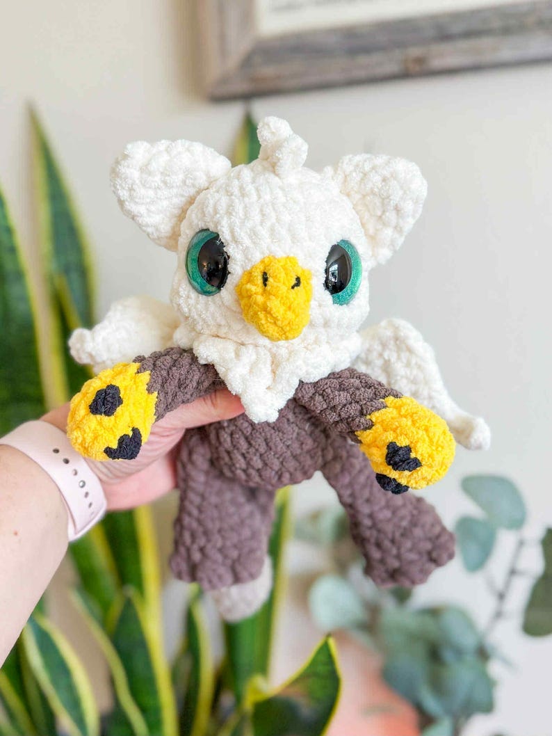 May include: A handmade crochet plush toy in the shape of a bird. The toy features a white head and wings, a yellow beak, and large green eyes. The body is brown with yellow and black accents on the feet. The plush is held by a person.