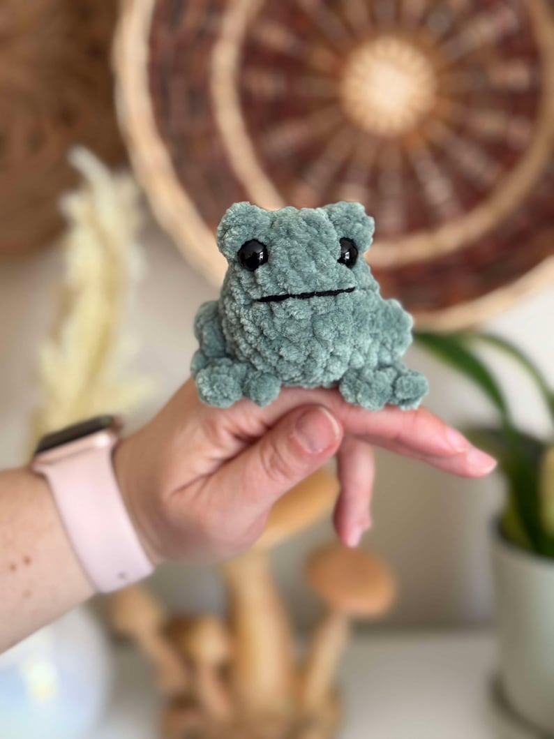 May include: A small, green crocheted frog with black eyes. The frog is sitting on a finger.