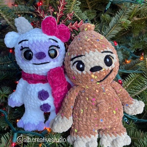 May include: Two crocheted plush toys, one white with purple accents and a pink bow, the other brown with colorful sprinkles, are displayed on a Christmas tree.