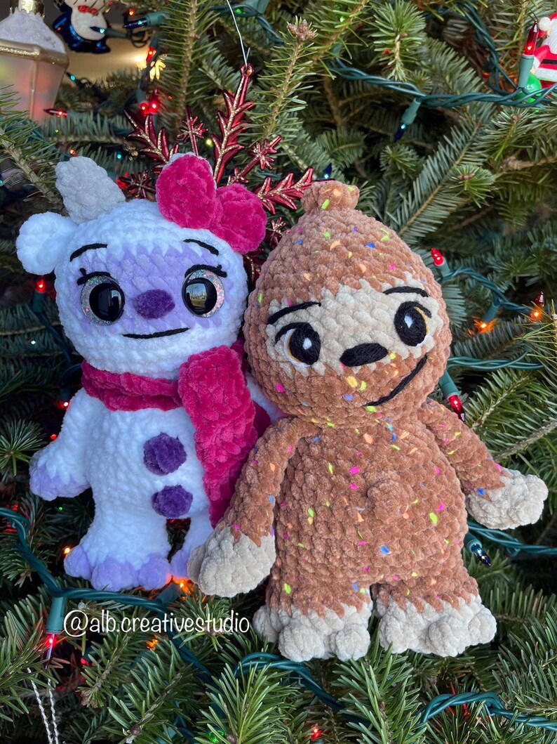 May include: Two crocheted plush toys, one white with purple accents and a pink bow, the other brown with colorful sprinkles, are displayed on a Christmas tree.