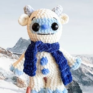 May include: White crocheted plush toy with blue and white accents, wearing a blue scarf. The toy has a friendly face with large black eyes and a blue nose.