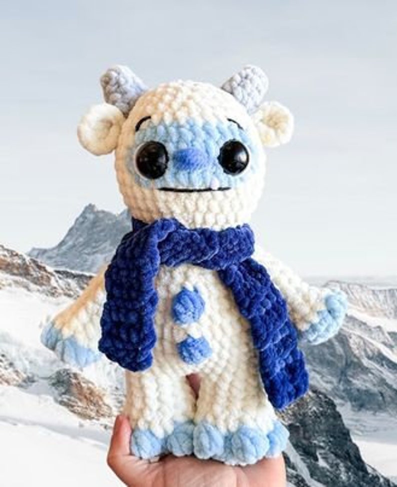 May include: White crocheted plush toy with blue and white accents, wearing a blue scarf. The toy has a friendly face with large black eyes and a blue nose.