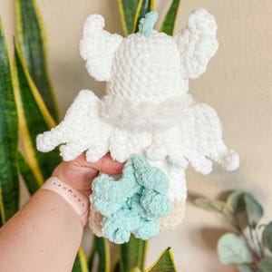 May include: A handmade white crochet angel doll with fluffy wings and a light blue accent. The doll has a soft, textured appearance and is held in a person's hand. The angel's wings are spread out, and the doll has a light beige base.