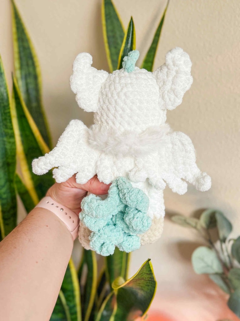 May include: A handmade white crochet angel doll with fluffy wings and a light blue accent. The doll has a soft, textured appearance and is held in a person's hand. The angel's wings are spread out, and the doll has a light beige base.