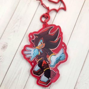 May include: Red acrylic keychain featuring a cartoon character with black and red fur, wearing a black jacket and white gloves. The keychain has a red bat-shaped top and a silver-colored ring.
