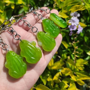 May include: A collection of green frog-shaped charms attached to silver-colored chains. The charms are a vibrant green color and have a glossy finish. The background features green and yellow foliage with small purple flowers.