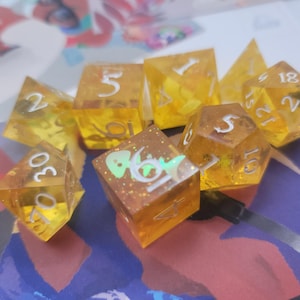 May include: A set of eight yellow dice with white numbers. The dice are translucent and have a glittery finish. The numbers on the dice are 1, 2, 3, 4, 5, 6, 18, and 20.