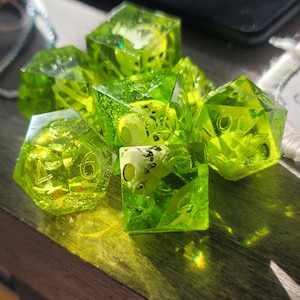 May include: A set of six green dice with white numbers. The dice are made of clear plastic and have a glittery finish. The dice are scattered on a wooden surface.