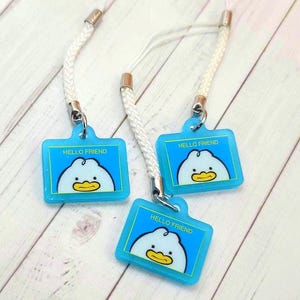 May include: Three blue Hello Friend charms with a white duck design and a white cord. The charms have a rectangular shape with rounded corners. The text "HELLO FRIEND" is printed on the top of the charm.
