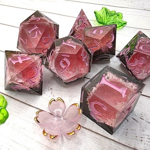 May include: A set of seven polyhedral dice made of clear resin with pink floral inclusions and light pink numbering. The set includes a d20, d12, d10, d8, d6, d4, and a percentile die, accompanied by a small, light pink, five-petaled flower charm.