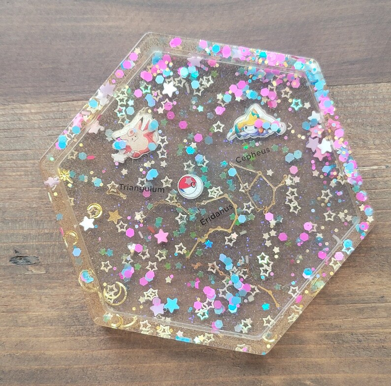 May include: A clear, hexagonal resin tray with embedded glitter and star-shaped confetti in pink, blue, and gold. The tray features constellation designs and cartoon character pins. Text includes "Triangulum", "Cepheus", and "Eridanus".