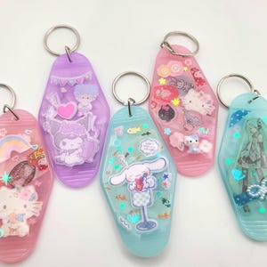May include: Five colorful keychains with silver key rings. The keychains are in shades of pink, purple, and blue, each featuring different cartoon characters and designs. The keychains have a retro motel key shape.